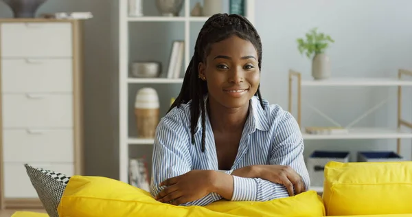 Portret atrakcyjnej młodej Afroamerykanki z ogonem warkoczyka opierającej się na żółtej sofie w salonie, patrzącej na bok i uśmiechającej się prosto do kamery. Wewnątrz — Zdjęcie stockowe