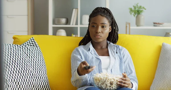 Ładna Afroamerykanka ogląda telewizję z dużą miską popcornu, zmienia kanały i siedzi na żółtej kanapie w nowoczesnym salonie. W pomieszczeniach — Zdjęcie stockowe