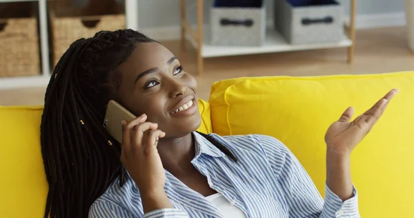 Muotokuva nuoresta afroamerikkalaisesta naisesta, joka puhuu iloisesti puhelimessa istuessaan keltaisen limsan päällä kotona. Sisätilat — kuvapankkivalokuva