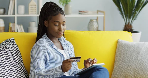 Mulher afro-americana bonita comprando on-line com um cartão de crédito no dispositivo tablet enquanto sentado no sofá na sala de estar. Interior — Fotografia de Stock