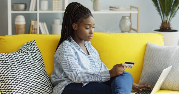 Houkutteleva nuori afroamerikkalainen nainen tekee ostoksia verkossa syöttäessään luottokortin numeron kannettavan tietokoneen kotona. Sisällä — kuvapankkivalokuva