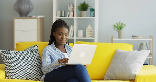 Kaunis nuori afroamerikkalainen nainen istuu sohvalla olohuoneessa ja käyttää kannettavaa tietokonetta, sitten hänen puhelimensa soi ja hän puhuu. Sisätilat — kuvapankkivalokuva
