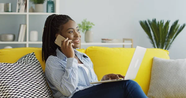 Pretty African American woman calling and speaking on the phone while sitting on the couch in the living room with a laptop computer on her knees. Inside