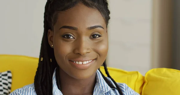 Sarı kanepede, saçı örgülü ve kameraya poz veren güzel Afro-Amerikan genç bayanı yakından çek. Kapalı