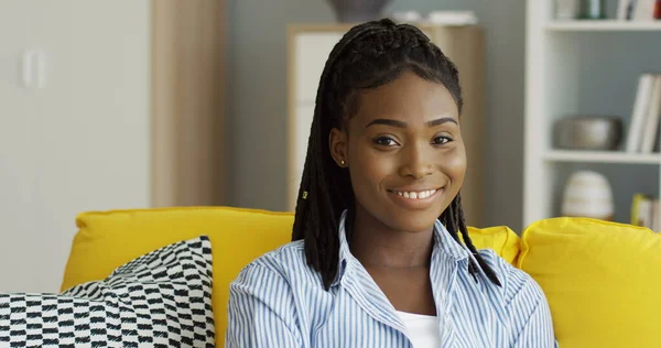 Portaali viehättävästä afroamerikkalaisesta nuoresta naisesta, jolla on saparot hymyilemässä kameralle istuessaan keltaisella sohvalla. Sisätilat — kuvapankkivalokuva