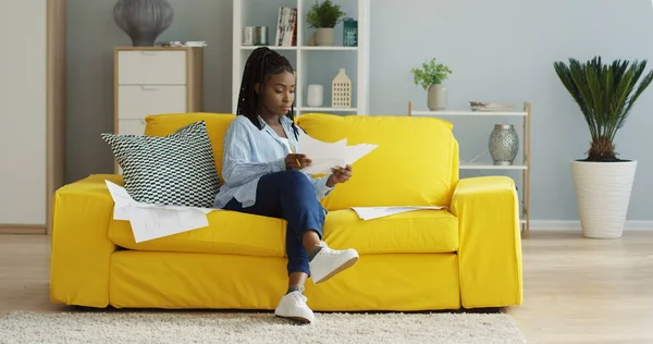 Spor elbiseli genç Afrikalı Amerikalı kadın oturma odasındaki sarı koltukta oturuyor ve belgelerle çalışıyor. İçeride.
