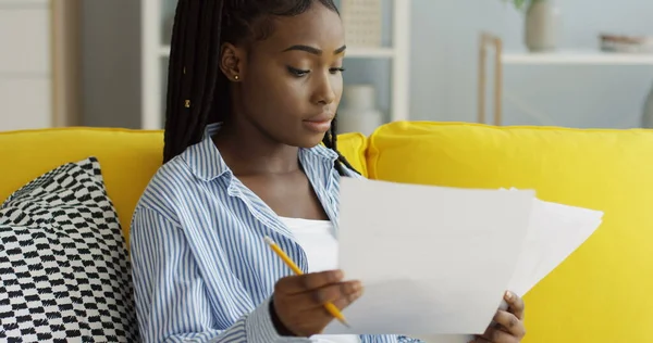 Close up da mulher afro-americana segurando folhas de papel e lápis nas mãos e verificando documentos no sofá em casa. Interior — Fotografia de Stock