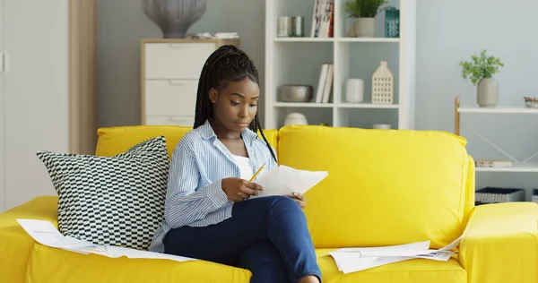 Kaunis nuori afroamerikkalainen nainen lukee joitakin asiakirjoja työskennellessään keltaisella sohvalla viihtyisässä olohuoneessa. Sisätilat — kuvapankkivalokuva