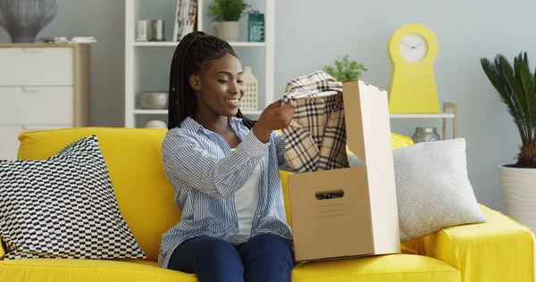 Muotokuva nuoresta afroamerikkalaisesta naisesta hakemassa postipakettia, avaamassa laatikkoa, ottamassa kirjavaa paitaa ja tyytymässä siihen. Sisätilat — kuvapankkivalokuva