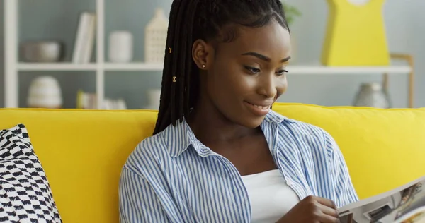 Evdeki sarı koltukta oturan ve dergi okurken dinlenen güzel Afrikalı Amerikalı kadının portresi. Kapalı