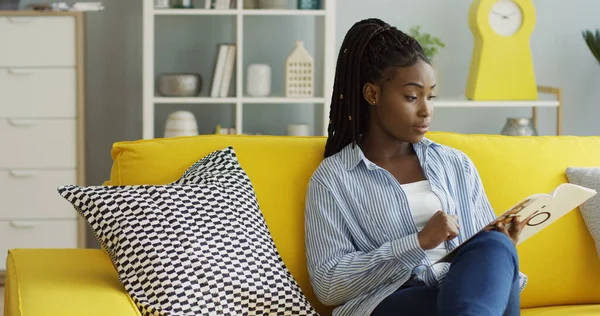 Afrikkalainen amerikkalainen nainen rento vaatteet flipping sivut lehden lukiessaan sitä sohvalla un moderni olohuone. Sisällä — kuvapankkivalokuva