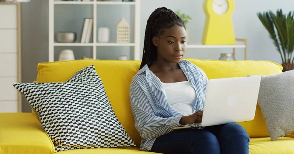 Nuori kaunis afroamerikkalainen nainen, jolla on häntä, joka työskentelee kannettavalla tietokoneella, vierittää ja teipata näppäimistöllä, kun hän istuu sohvalla modernissa olohuoneessa. Sisällä — kuvapankkivalokuva