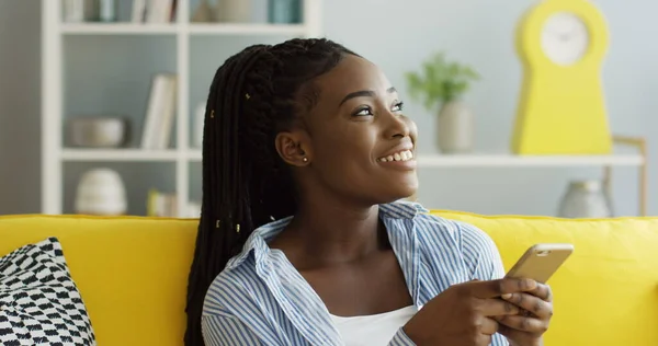 Nær den unge, attraktive afroamerikanske kvinnen med hale som ruller og teiper seg på smarttelefonen mens hun sitter på sofaen i den koselige stuen. Innendørs – stockfoto