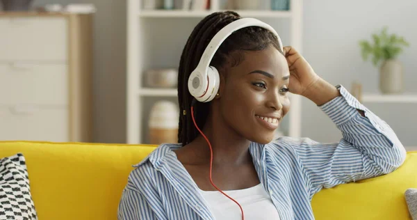 Portrett av den unge, smilte afroamerikanske kvinnen med hodetelefoner mens hun lyttet til musikken på den gule sofaen hjemme. Innendørs – stockfoto