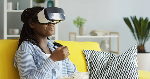 Портрет молодої афро-американської жінки, яка сидить на дивані і має VR навушники, спостерігає, як щось їсть попкорн і посміхається. У приміщенні Стокова Картинка