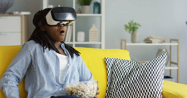 Portrait shot of the African American woman with pigtails sitting on the sofa during VR headset, watching something scary while eating popcorn and being afraid. Inside Stock Photo