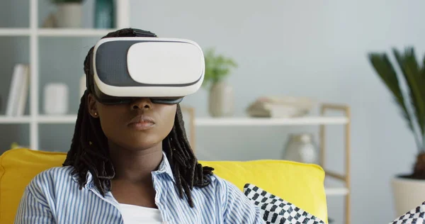 Nær den afroamerikanske unge kvinnen i VR-briller som ser på noe mens hun har hodetelefoner og spiser popkorn. Innendørs stockbilde