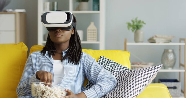 Portrait of the African American woman with pigtails and with bowl full of popcorn having VR headset, watching something that being scary or shocking in the living room. Indoors Royalty Free Stock Photos