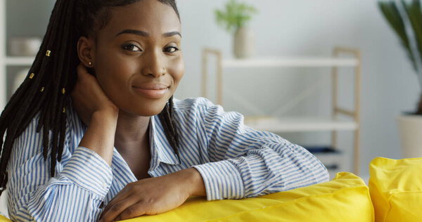 Charming African American young woman in the blue shirt sitting on the yellow couch and thinking, then smiling at the camera. Indoors Stock Picture