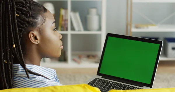 Vedere peste umăr pe tânăra afro-americană cu cozi așezate în fața laptopului deschis cu ecran verde și gândire. Cheia cromatică. De aproape. Interior fotografii de stoc fără drepturi de autor