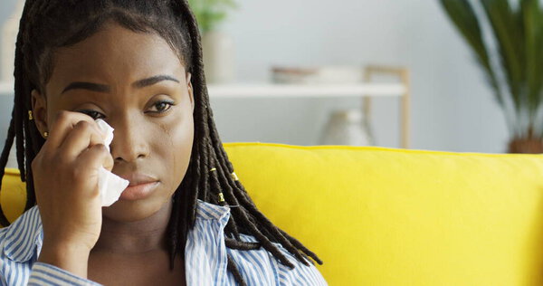 Close up of the sad African American young woman crying while watching movie on the yellow sofa at home and wiping her tears with a napkin. Indoors Stock Photo
