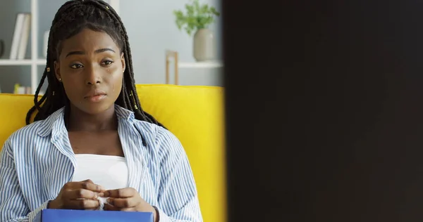 Upset African American woman in the blue shirt and with pigtails sitting on the yellow couch in the living room in front of the TV screen and crying with a napkin in a hand. Inside Stock Photo