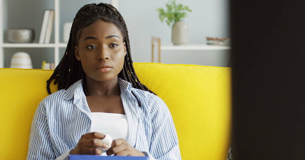 Attractive African American young woman resting on the yellow sofa at home and watching TV, crying because of the sad film. Indoor Stock Photo