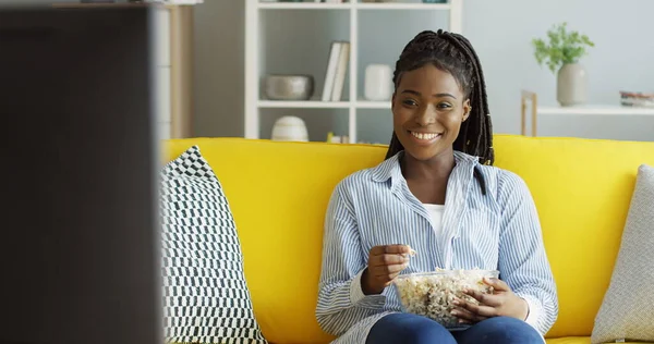 Frumoasa femeie afro-americană în cămașa albastră mănâncă popcorn în timp ce se uită la un film de comedie la televizor și râde pe canapeaua galbenă acasă. Inauntru Fotografie de stoc