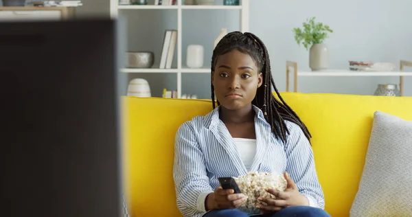 Portretul unei tinere afro-americane care mananca popcorn si se uita la televizor pe canapeaua galbena din sufrageria moderna. Interior Imagine de stoc