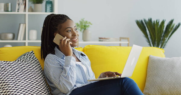 Pretty African American woman calling and speaking on the phone while sitting on the couch in the living room with a laptop computer on her knees. Inside Stock Image