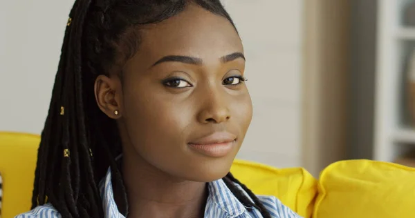 Close-up van de mooie Afro-Amerikaanse jonge vrouw met een staart van vlechten kijkend en poserend voor de camera op de gele bank. Binnenshuis Rechtenvrije Stockafbeeldingen