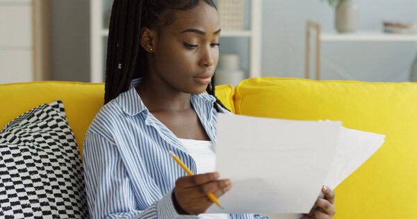 Close up of the African American woman holding paper sheets and pencil in hands and checking documents on the couch at home. Indoor Royalty Free Stock Photos