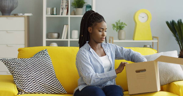 Young African American woman getting postal parcel, opening a box, getting out motley shirt and being unsatisfied. Inside Stock Picture