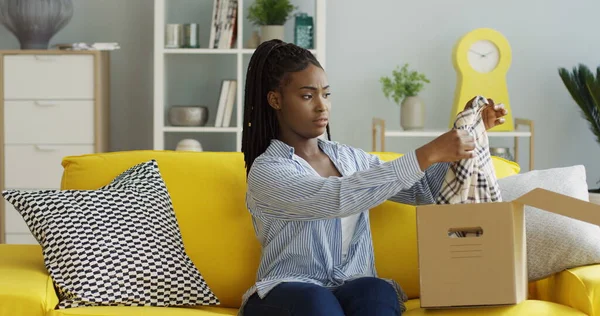 Jeune femme afro-américaine recevant un colis postal, ouvrant une boîte, sortant une chemise bigarrée et insatisfaite. À l'intérieur Photo De Stock