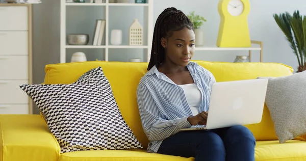 En ung, afroamerikansk kvinne som skriver på PC-en og lager grimaser på grunn av noe hun leser. Hjemme. Innendørs stockbilde