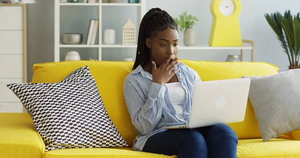 Досить афроамериканська молода жінка, яка друкує смс на ноутбуці комп'ютера на колінах і робить вау-обличчя через щось, що вона читає. Вдома. У приміщенні Ліцензійні Стокові Фото