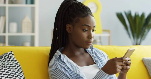 Nær den unge, attraktive afroamerikanske kvinnen med hale som ruller og teiper seg på smarttelefonen mens hun sitter på sofaen i den koselige stuen. Innendørs stockbilde