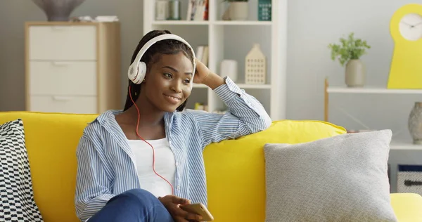 Ung, lykkelig afroamerikansk kvinne med store hodetelefoner som nyter musikken hun holder i smarttelefonen mens hun hviler på den gule sofaen hjemme. Innvendig stockbilde