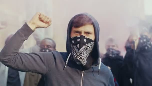Zbliżenie białego przystojnego młodzieńca w kapturze i szalik na twarzy protestujących i krzyczących sloganów w dymie i gazie na zewnątrz. Wieloetniczni faceci strajkujący na ulicy w USA. — Wideo stockowe