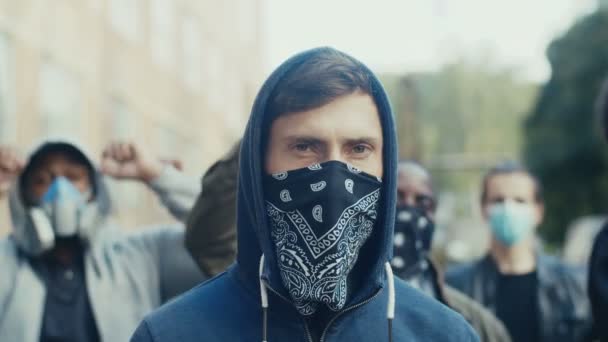 Retrato de un joven guapo hombre caucásico con capucha y bufanda en la máscara mirando a la cámara mientras está de pie en la multitud de pretesters. Primer plano de cara masculina enmascarada al aire libre en huelga . — Vídeos de Stock