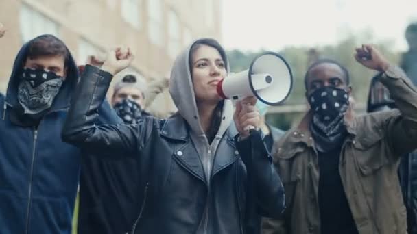 Caucasian young woman in hood screaming mottos in megaphone. Mixed-races male and female crowd of protesters shouting slogans and protesting. Girl leading at manifestation for equality. — Stock Video