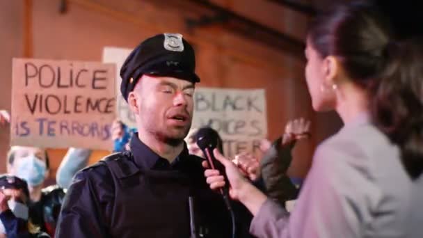 Correspondente caucasiana entrevistando policiais na rua à noite, enquanto as pessoas protestam pelos direitos afro-americanos e contra a violência policial. Entrevista de policial por mulher para notícias nos EUA . — Vídeo de Stock