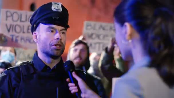 Beyaz kadın muhabir gece sokakta polisle röportaj yaparken Afro-Amerikan haklarını ve polis şiddetini protesto eden insanlar. Amerika 'da haber için kadın kadın polis röportajı. Arka.. — Stok video