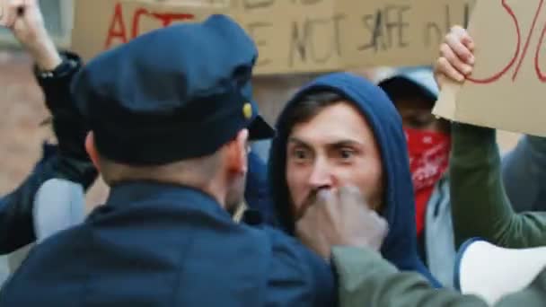 Кавказький молодий чоловік протестує в капюшоні, кричачи і нападаючи на поліцейського на знак протесту проти насильства і расизму. Поліцейські використовують силу, щоб заспокоїти чоловіків-бунтівників. Бої поліції під час бунту.. — стокове відео