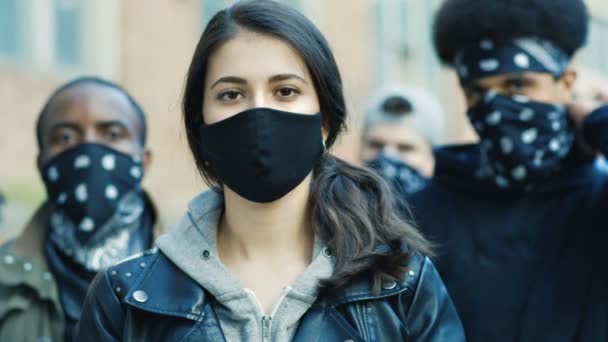 Primer plano de la joven caucásica hermosa mujer en la máscara mirando directamente a la cámara al aire libre en la calle motín. Retrato de una chica con afroamericanos masculinos en el fondo. Manifestante femenina en manifestación . — Vídeo de stock