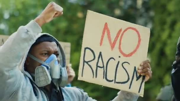 Hombre joven afroamericano manifestante con capucha y máscara respiratoria sosteniendo afiche Sin racismo y protestando contra la violencia policial. Hombre gritando y gritando en la calle motín . — Vídeos de Stock