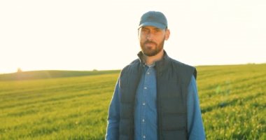 Şapkalı ve kot pantolonlu yakışıklı beyaz adamın yeşil alanda kameraya neşeyle gülümseyip baş parmağını verdiği portre resmi. Yazın açık havada gülümseyen erkek çiftçi..