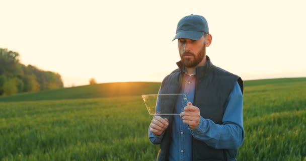 Kaukázusi fiatal vonzó férfi kalapban áll zöld mezőn, üvegre koppint, mint egy futurisztikus eszköz képernyőjén. Képernyőérzékelés. Jövőbeli technológia. Férfi fermer átlátszó számítógéppel. — Stock videók