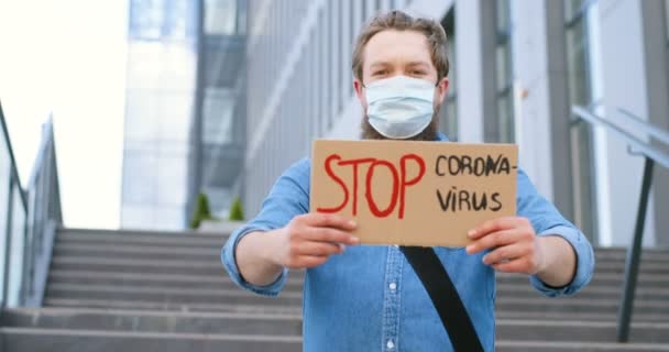 Retrato de un joven caucásico con máscara médica parado al aire libre en grandes escaleras y mostrando la mesa con palabras Stop Coronavirus. Junta de manifestantes masculina con protesta contra pandemia. — Vídeos de Stock