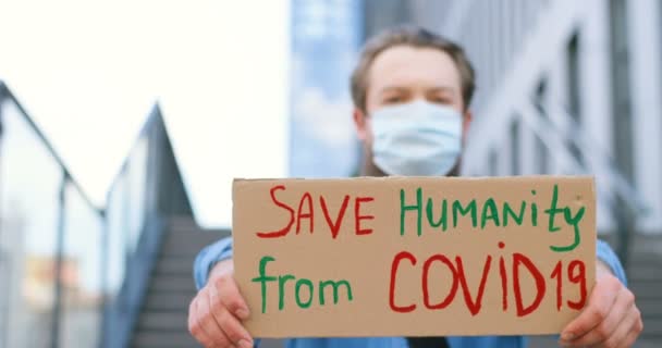 Close up of carton table with words Save Humanity from Covid 19 in hands of Caucasian young man in medical mask. Blurred male behind. Lonely protest outdoors. — Stock Video